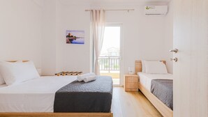 A hotel room with two beds, a wooden bench, a small bedside table, a wall-mounted air conditioner, and a window with a view of a building and a body of water. - Artistic Vibe by Rentalspro - Nikiti Halkidiki (Sithonia)
