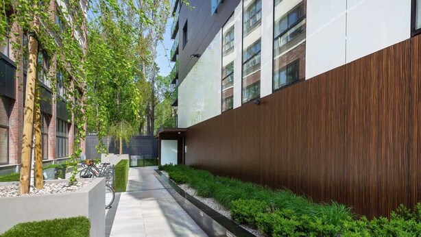 A modern building with a clean, straight pathway flanked by greenery and a wooden wall.