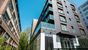 A modern multi-story building with glass balconies, a mix of brick and glass facade, and a clear blue sky in the background.
