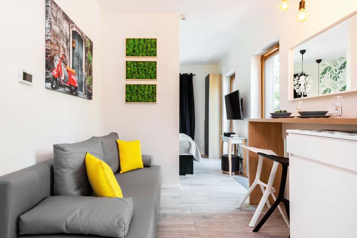 Un salon moderne avec un canapé gris et des coussins jaunes, un coin repas avec une table et des chaises blanches, et une cuisine dotée d’un comptoir en bois.