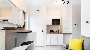 A modern kitchen with white cabinets, a black oven, and a coffee maker. There is a dining area with a white chair and a table. A gray sofa with a yellow pillow is in the living room.