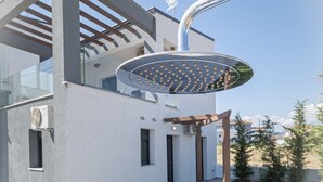 Eine moderne Außendusche mit Metallrahmen und kreisförmigem Design, an einem zeitgemäßen Gebäude mit weißer Fassade und einer Pergola.