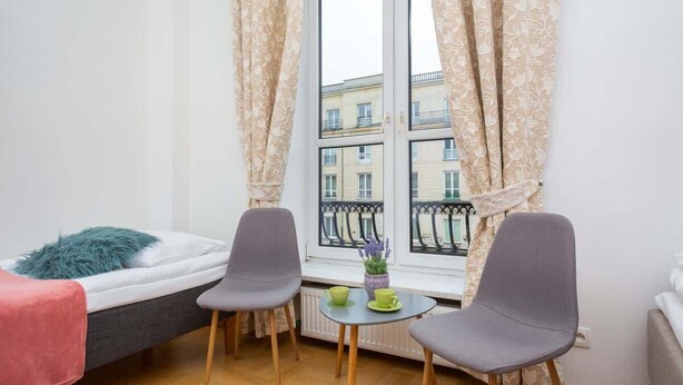 Una habitación pequeña y moderna con una cama, dos sillas, una mesita con tazas y una ventana con cortinas.