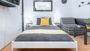 A modern bedroom with a bed, a black and white patterned armchair, a small table, a wall-mounted TV, a mirror, and a clock. - Cozy Studio in the Center | Close to Central Station (Warszawa)