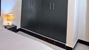 A bedroom with a large wardrobe, a bedside table with a lamp, and a bed with white bedding. - B&S Luxe Living (Kampala)