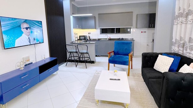 A modern living room with a flat-screen TV, a blue entertainment unit, a white coffee table, a black sofa, and a kitchen in the background. - B&S Luxe Living (Kampala)