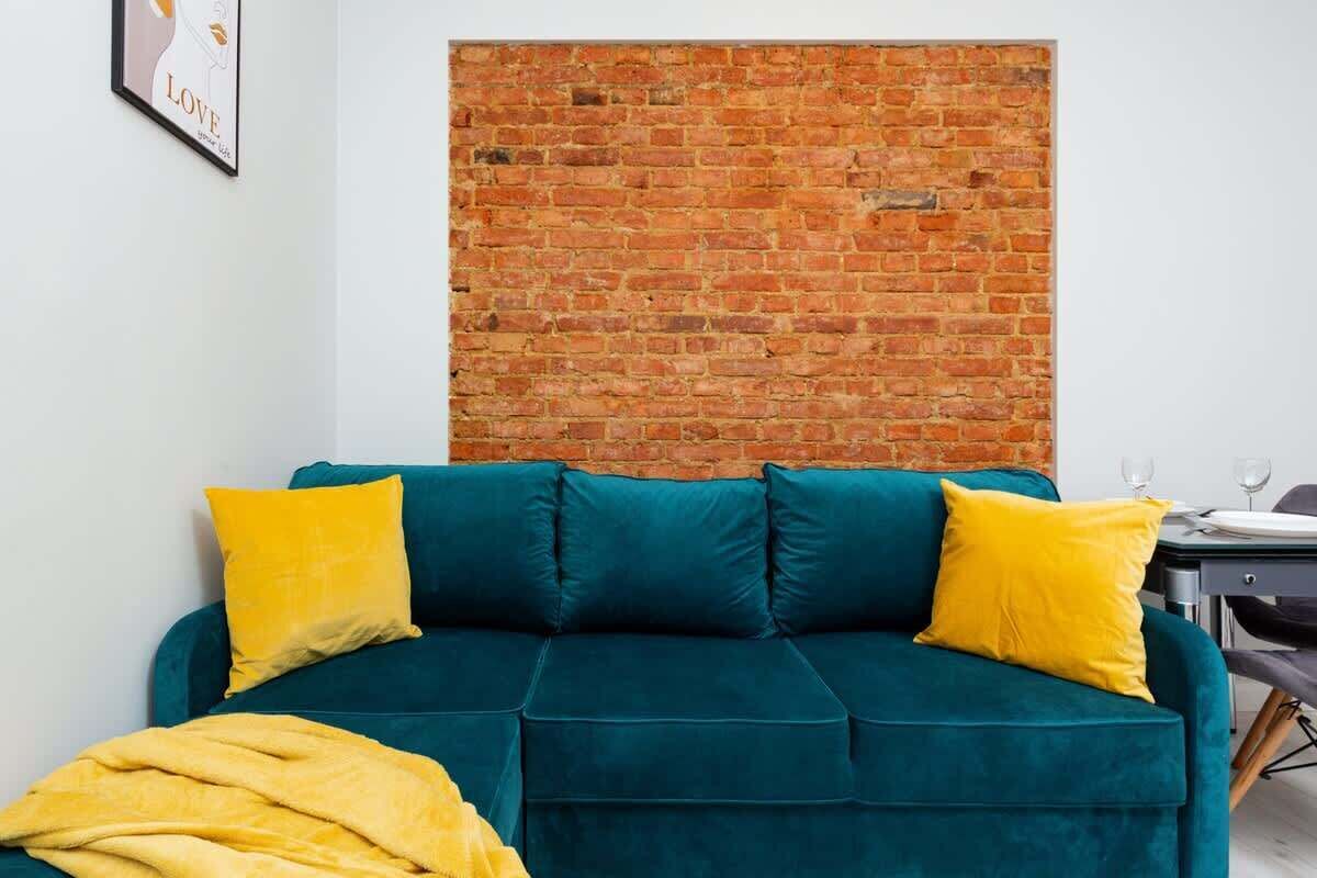 A teal sofa with yellow cushions, a brick wall, and a framed picture on the wall.