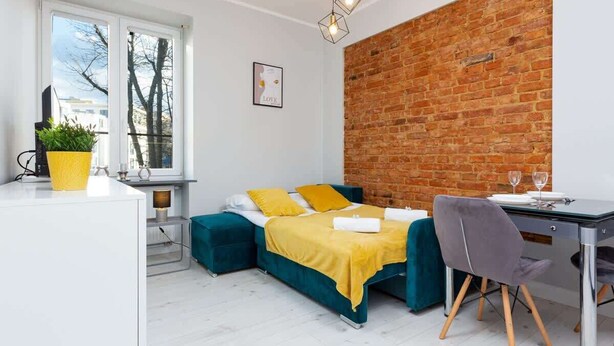 Ein modernes Zimmer mit Backsteinwand, einem Bett mit gelber Bettwäsche, einem Schreibtisch mit Stuhl und einem Fenster mit Blick auf Bäume.