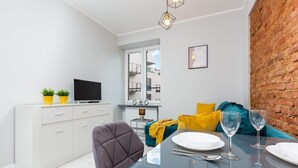 Ein gemütlicher Wohnraum mit Esstisch für zwei Personen, einer Couch mit Kissen, einem Fernseher auf einem weißen Schrank und einer Ziegelwand.