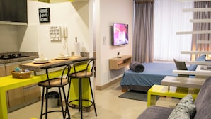 Ein kompakter Wohnraum mit Küchenzeile, einer Essecke mit zwei Barhocker, einem Bett mit blauer Bettdecke, einem an der Wand montierten Fernseher und einem Fenster mit Vorhängen.