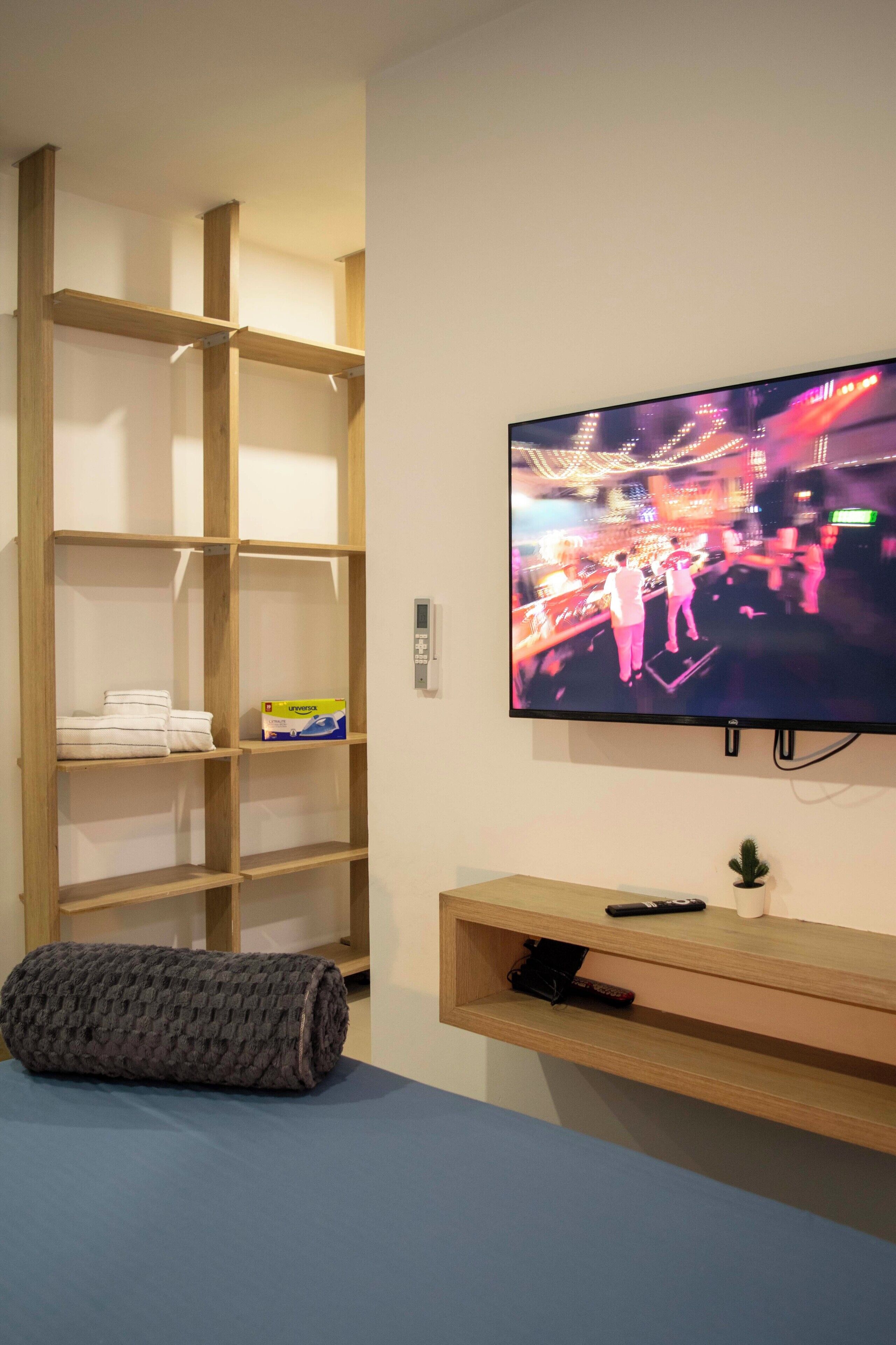 Een kamer met een aan de muur bevestigde televisie, een houten plank met handdoeken, een afstandsbediening en een kleine plant.