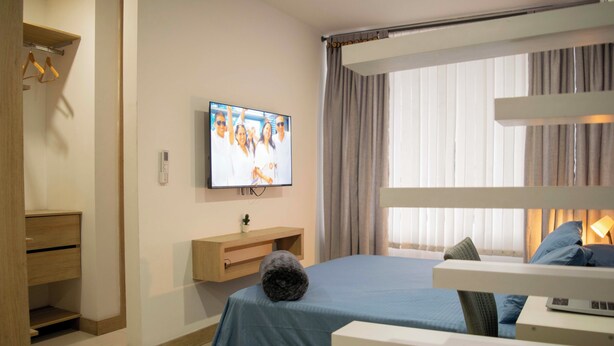 Ein Hotelzimmer mit Bett, einem an der Wand befestigten Fernseher, einem Nachttisch mit Lampe und einem Fenster mit Vorhängen.