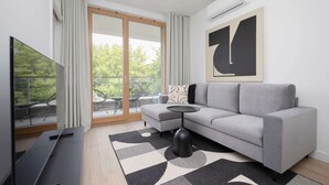 A modern living room with a grey sofa, a black coffee table, a flat-screen TV, and a large window with a view of greenery.
