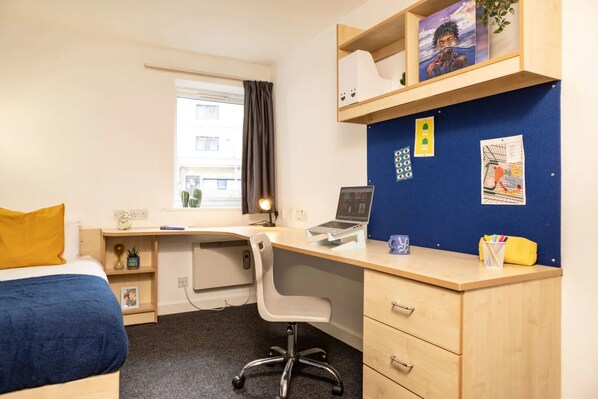 A hotel room with a bed, a desk with a laptop, a chair, a window with curtains, and a bulletin board with various items pinned to it.