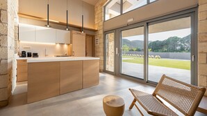 A modern kitchen with wooden cabinets, a central island, pendant lighting, and a large sliding glass door leading to a patio with a view of a green landscape.