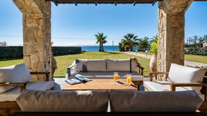 Ein von Steinbögen geschützter Außenbereich mit Sitzgelegenheit, einem Holztisch, Kissen und Blick auf das Meer.