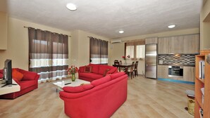 Una sala de estar con un sofá rojo, una mesa de centro y un televisor. Hay una zona de cocina con un refrigerador, estufa y una mesa de comedor.