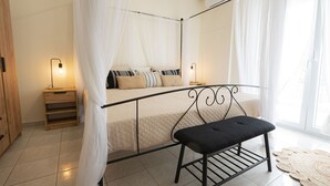 Een slaapkamer met een hemelbed, nachtkastjes, een bankje en een groot raam met dunne gordijnen.