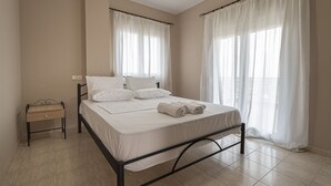 Een net opgemaakt bed met witte lakens en handdoeken, een nachtkastje en een raam met dunne gordijnen.
