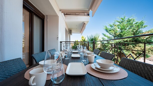 A balcony with a table set for dining, featuring clear glassware and plates, and a view of greenery and a building in the background.