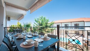 A balcony with a table set for dining, overlooking a swimming pool and a building with a red-tiled roof.