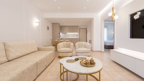 A modern living room with a beige sofa, two armchairs, a round coffee table with decorative items, and a kitchen in the background. - Les Maisons Deluxe Superior by Rentalspro (Kassandra)