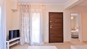 A hotel room with a wooden door, a television on a stand, a white sofa, and a large window with sheer curtains. - Fotinia Beach House Ground Floor by Rentalspro (Nea Propontida)