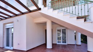 A spacious outdoor area with a tiled floor, a staircase with a green railing, and a glass door leading to a covered balcony. - Fotinia Beach House Ground Floor by Rentalspro (Nea Propontida)