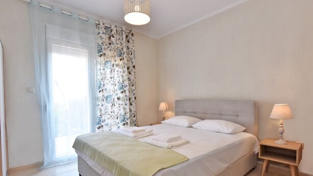 A neatly made bed with white linens and a green throw blanket, two bedside lamps, and a window with floral curtains. - Fotinia Beach House Ground Floor by Rentalspro (Nea Propontida)
