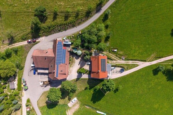 An aerial view of a building with solar panels, surrounded by green fields and a winding road.
