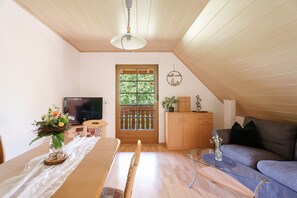 A cozy living room with a wooden ceiling, a dining table with a white tablecloth, a sofa, and a television.