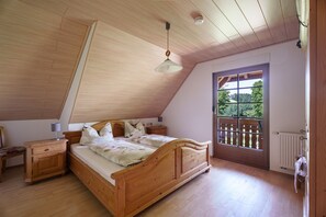 Ein Schlafzimmer mit schrägen Wänden, einem hölzernen Bett, einem Nachttisch, einer Kommode und einem Fenster mit Blick ins Grüne.
