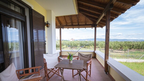 A covered outdoor area with a table and chairs, a potted plant, and a view of a distant building and landscape. - Casa Natura Rodanthi by Rentals Pro (Nea Propontida)