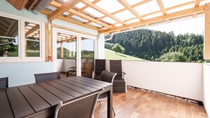 En overdækket terrasse med et bord og stole, udsigt til en skovklædt bakke, og et træloft.