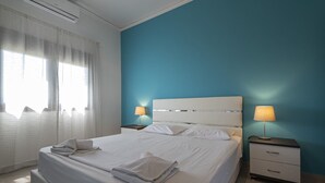 A neatly made bed with white linens and a folded towel, two bedside tables with lamps, a window with sheer curtains, and a blue accent wall. - Villa Elva by Rentalspro (Sithonia)