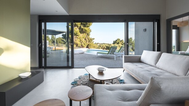 A modern living room with a sofa, coffee table, and a view of a pool and outdoor seating area through large sliding doors. - Theeta Elia Villa - Metamorfosi Halkidiki (Sithonia)