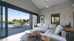 A modern living room with a large sofa, a coffee table, and a view of a pool and greenery outside. - Theeta Elia Villa - Metamorfosi Halkidiki (Sithonia)