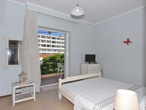 Ein Schlafzimmer mit einem Bett, einem Schreibtisch mit Computer, einem kleinen Tisch mit Lampe und einem Fenster mit Vorhängen.