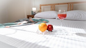 Un lit bien fait, sur lequel repose un plateau en verre contenant une orange et des fraises, un verre d’une boisson rouge et une lampe de chevet.