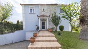 Une maison à deux étages avec une façade en pierre, un toit en tuiles et une entrée à l’avant. Des marches mènent à la porte, et des plantes en pot sont disposées le long du chemin.
