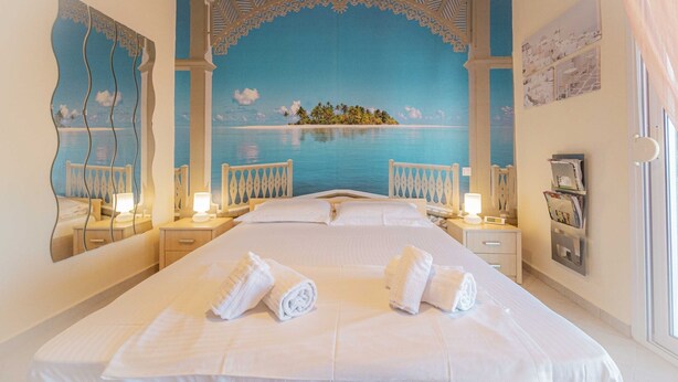 A large bed with white linens and two rolled towels, a nightstand with a lamp, a wall-mounted shelf with magazines, and a large window with curtains. - Eternal Blue by Rentalspro-hanioti Halkidiki (Kassandra)
