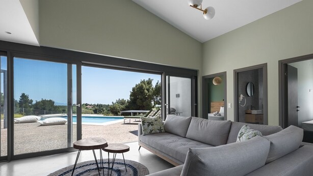 A modern living room with a large sofa, a coffee table, and a view of a swimming pool and greenery outside. - Theeta Pefko Villa - Metamorfosi Halkidiki (Sithonia)
