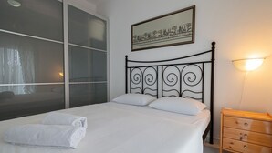 A neatly made bed with white linens and two rolled towels, a wooden nightstand with drawers, a wall-mounted picture, and a window with curtains. - Magdalini's Seaview House - Pefkohori (Kassandra)