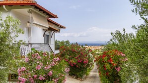 A white building with a red-tiled roof and a balcony. A pathway lined with pink and red flowers leads to a view of a coastal town. - Magdalini's Seaview House - Pefkohori (Kassandra)