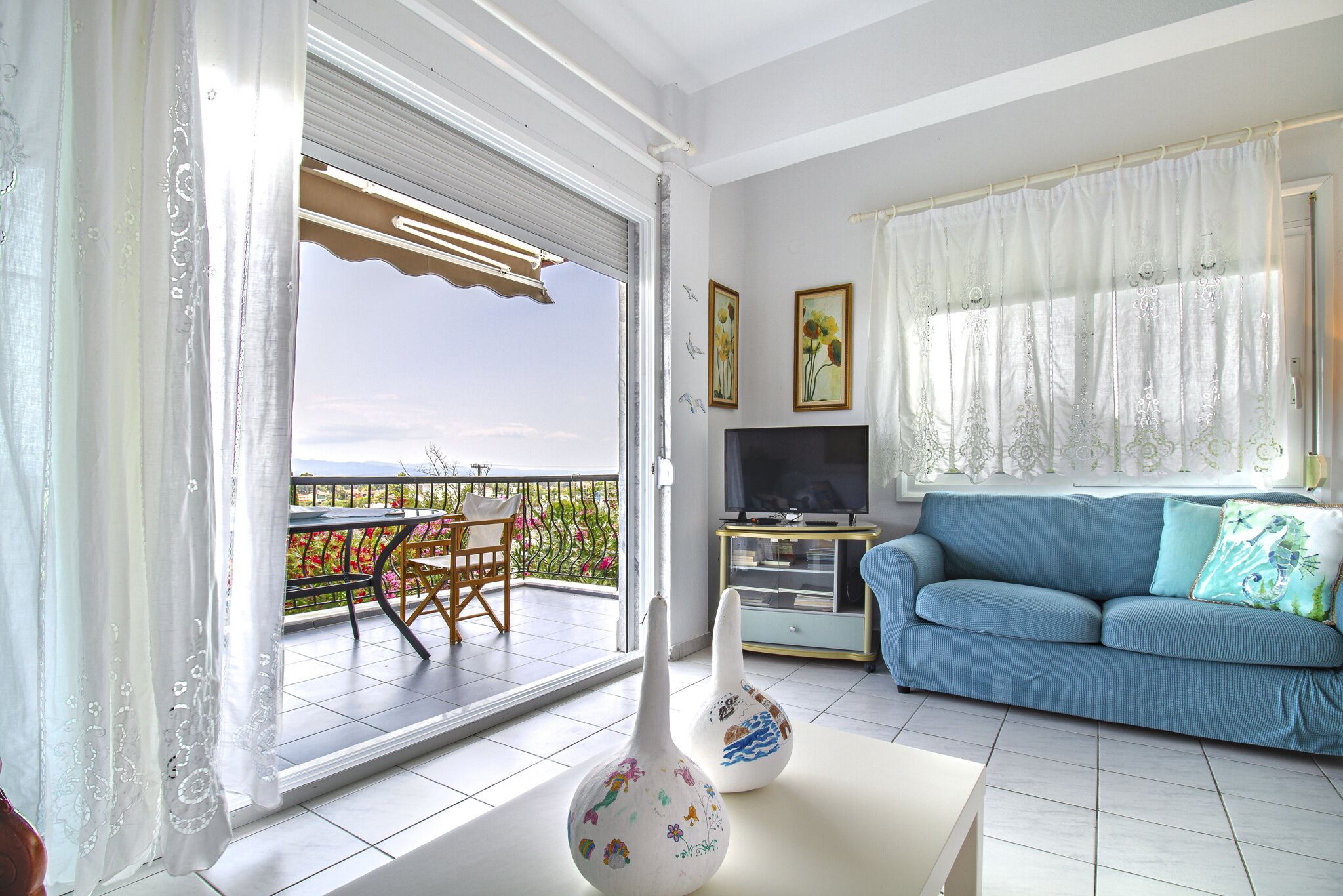 Una sala de estar con sofá, televisión, jarrones decorativos y un balcón con mesa y sillas.