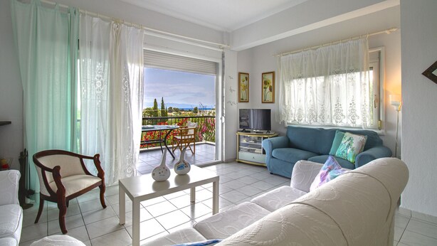 A living room with a sofa, a chair, a coffee table, and a TV. There is a balcony with a view of the city. - Magdalini's Seaview House - Pefkohori (Kassandra)