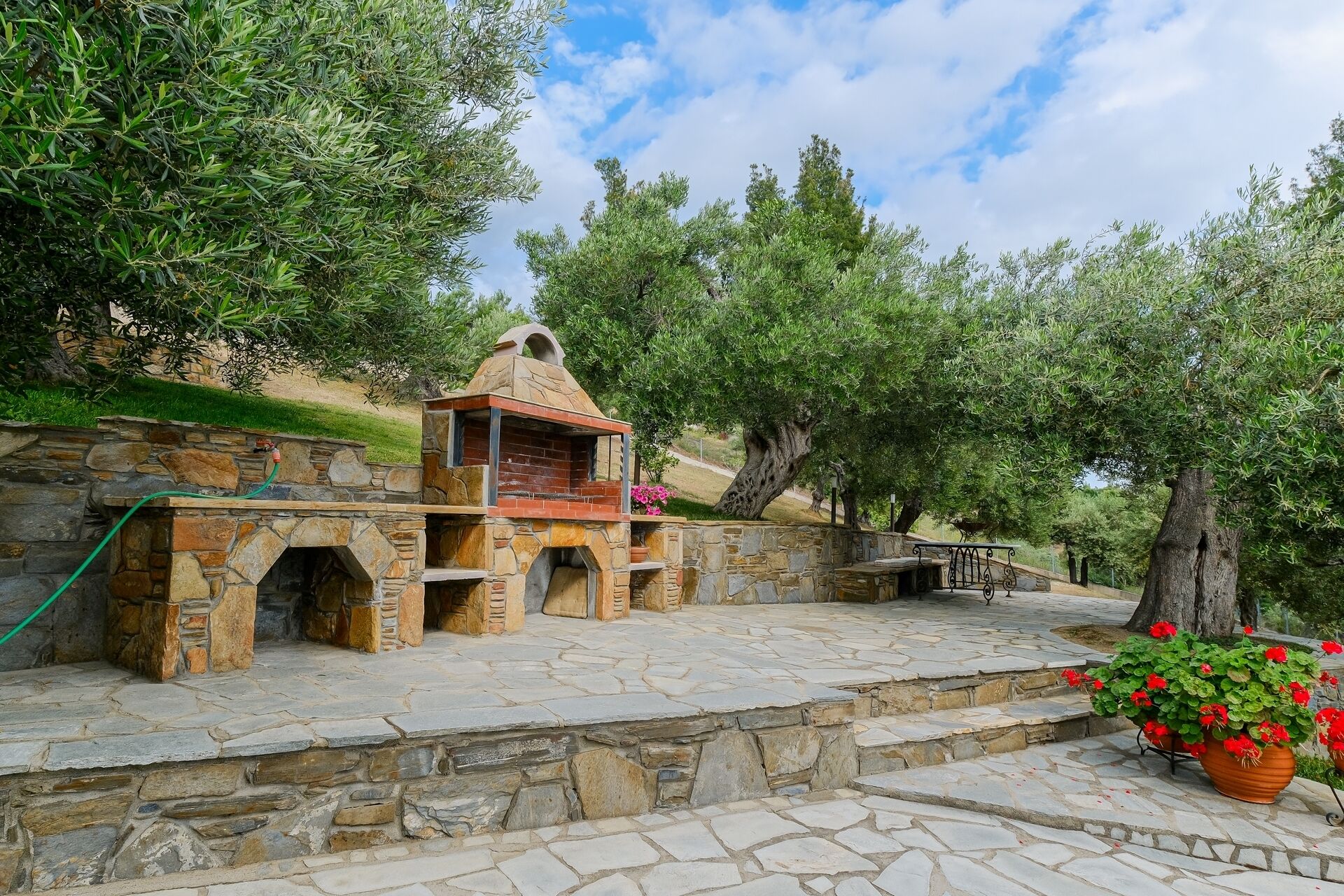 A stone-built outdoor area with a barbecue setup, stone steps, and a potted plant with red flowers.