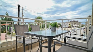 A balcony with a glass table and chairs, a potted plant, and a view of the street and buildings. - Tommy Seafront No 2 by Rentalspro - Nea Flogita (Nea Propontida)