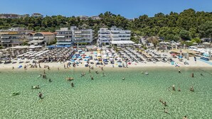 A beach with clear water, umbrellas, and people. There are buildings in the background.