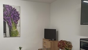 Un salon moderne avec une table à manger en bois, un téléviseur à écran plat posé sur un meuble en bois, une petite table d’appoint blanche agrémentée d’une plante, et un tableau représentant des fleurs violettes accroché au mur.